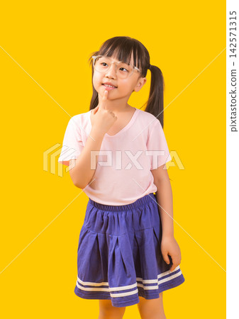 A cute little Asian girl puts her hand to her mouth, looks up at the side with a thoughtful expression, against a yellow studio background. A cute little Asian girl puts her hand to her mouth, looks up at the side with a thoughtful expression, against a yellow studio background. 142571315
