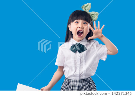 Expressive elementary age schoolgirl shouting with open mouth and waving hand against blue background. Expressive elementary age schoolgirl shouting with open mouth and waving hand against blue background. 142571345