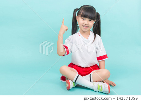 Smiling little girl in sports uniform sitting cross-legged and pointing finger up at copy space. Smiling little girl in sports uniform sitting cross-legged and pointing finger up at copy space. 142571359