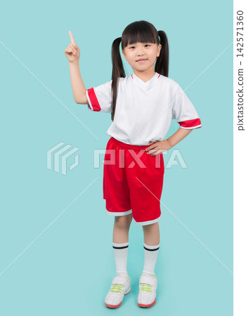 Cheerful little girl in red and white sports uniform standing and pointing finger up in studio Cheerful little girl in red and white sports uniform standing and pointing finger up in studio 142571360