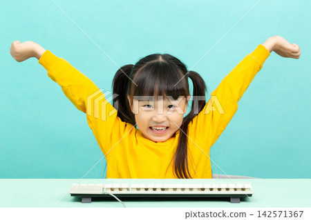 Joyful little girl raising hands in celebration behind a computer keyboard after achieving success. Joyful little girl raising hands in celebration behind a computer keyboard after achieving success. 142571367