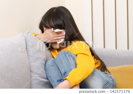 Sad little girl wiping her tears with a tissue while sitting on a living room sofa. 142571379