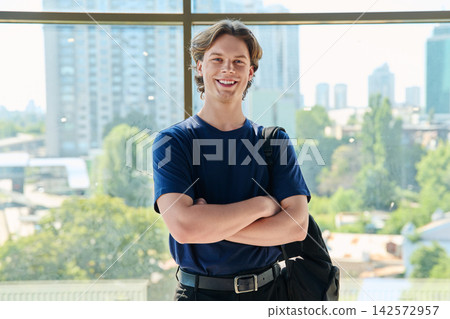 Smiling young male student standing with backpack and crossed arms near window 142572957