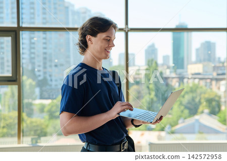 Smiling young male student holding laptop and typing near big window with city view Smiling young male student holding laptop and typing near big window with city view 142572958
