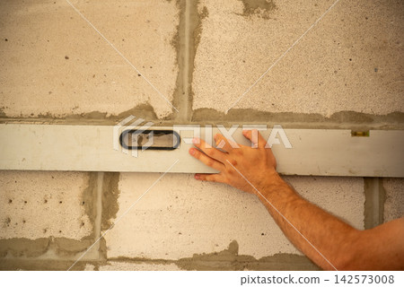 Hand holding a spirit level against a freshly laid concrete block wall, ensuring precision and accuracy during the building and construction process for a new home or renovation project Hand holding a spirit level against a freshly laid concrete block wall, ensuring precision and accuracy during the building and construction process for a new home or renovation project 142573008
