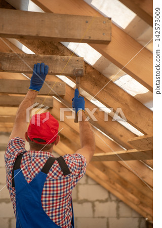 Builder in work clothes and gloves installing wooden roof truss beams with a hammer inside an unfinished house, framing the attic and creating a new home structure Builder in work clothes and gloves installing wooden roof truss beams with a hammer inside an unfinished house, framing the attic and creating a new home structure 142573009
