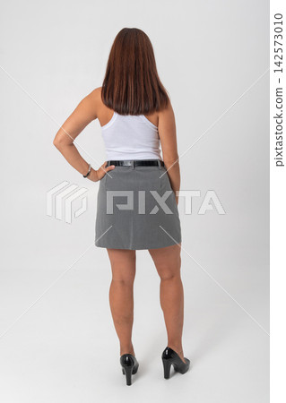 Woman standing with hand on hip, showing back, wearing a white tank top, grey skirt, and black high heels, indicating confidence and professional style 142573010
