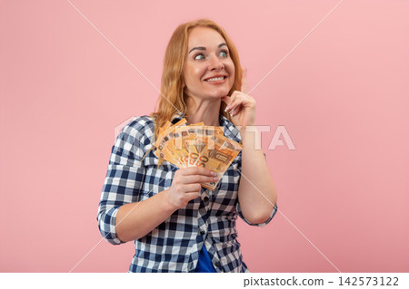 Smiling woman holding a fan of 50 euro banknotes, looking up and thoughtfully planning her finances, savings, and future investments on a pink background Smiling woman holding a fan of 50 euro banknotes, looking up and thoughtfully planning her finances, savings, and future investments on a pink background 142573122