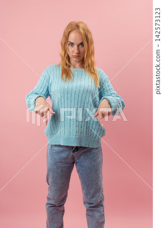 Young woman in a light blue sweater and jeans stands against a pink background, pointing her fingers downwards with a stern, concentrated facial expression, indicating direction or emphasis Young woman in a light blue sweater and jeans stands against a pink background, pointing her fingers downwards with a stern, concentrated facial expression, indicating direction or emphasis 142573123