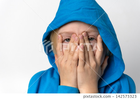 Young child wearing a blue hoodie covering face with both hands, looking through spread fingers with a shy or fearful expression against a plain white background Young child wearing a blue hoodie covering face with both hands, looking through spread fingers with a shy or fearful expression against a plain white background 142573323