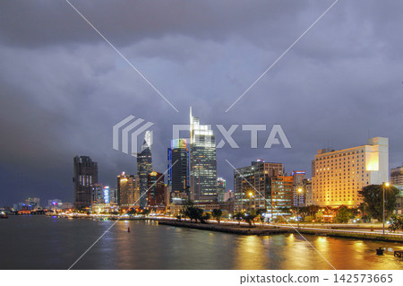 Wide nighttime cityscape showing illuminated modern towers, riverbank buildings, calm reflections on water, and dramatic storm clouds above busy downtown lights along waterfront, with skyline, city Wide nighttime cityscape showing illuminated modern towers, riverbank buildings, calm reflections on water, and dramatic storm clouds above busy downtown lights along waterfront, with skyline, city 142573665