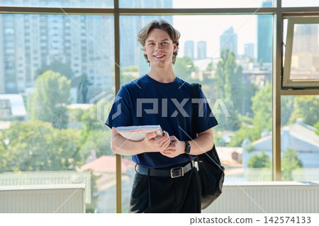 Young male student holding copybook standing looking at camera near window in campus Young male student holding copybook standing looking at camera near window in campus 142574133