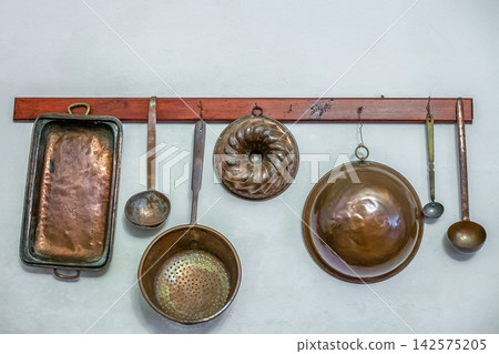 Vintage kitchen utensils hanging on wall in kitchen. Metal old kitchen appliances, baking molds, colander, ladles Vintage kitchen utensils hanging on wall in kitchen. Metal old kitchen appliances, baking molds, colander, ladles 142575205