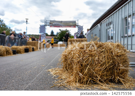 Hay bale marks safety barrier beside soap box racing course in Germany, with finish gate and spectators blurred behind. Community event combines speed, competition, tradition and summer atmosphere Hay bale marks safety barrier beside soap box racing course in Germany, with finish gate and spectators blurred behind. Community event combines speed, competition, tradition and summer atmosphere 142575556