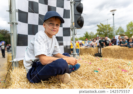 Boy watches soap box racing from hay bales beside checkered flag, enjoying outdoor community event in Germany. Young spectator rests near finish area while crowd gathers around downhill race course Boy watches soap box racing from hay bales beside checkered flag, enjoying outdoor community event in Germany. Young spectator rests near finish area while crowd gathers around downhill race course 142575557