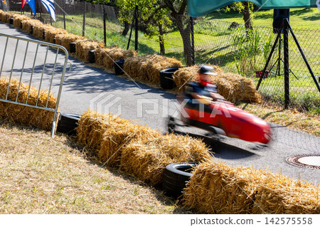 Soap box racer speeds through curved street course in Germany, captured with motion blur beside hay bales and tire barriers. Summer race conveys speed, competition, excitement and festival atmosphere Soap box racer speeds through curved street course in Germany, captured with motion blur beside hay bales and tire barriers. Summer race conveys speed, competition, excitement and festival atmosphere 142575558