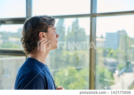Pensive young man looking out large window with city view in profile 142575606