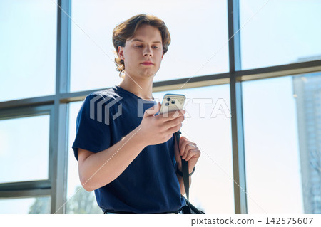 Young male student using smartphone with backpack standing near big window 142575607