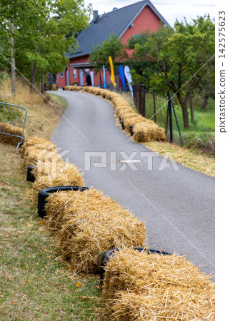 Empty soap box racing course winds downhill through rural German village, lined with hay bales and tires for safety. Curved asphalt track awaits racers during outdoor local community competition Empty soap box racing course winds downhill through rural German village, lined with hay bales and tires for safety. Curved asphalt track awaits racers during outdoor local community competition 142575623