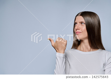 smiling young woman pointing thumb and looking aside at copy space on grey background 142575643