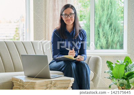 Smiling businesswoman holding pen and notebook looking at camera at cozy home office Smiling businesswoman holding pen and notebook looking at camera at cozy home office 142575645