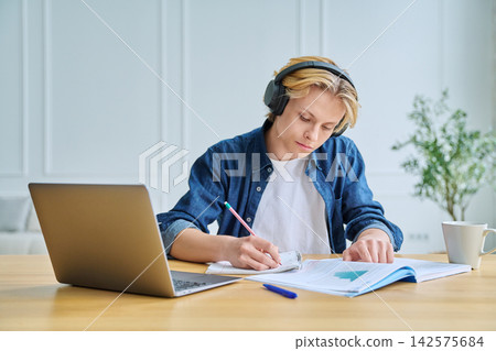 Teenage student in headphones writing at desk indoor Teenage student in headphones writing at desk indoor 142575684