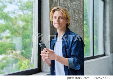 Smiling teenage student holding smartphone near window Smiling teenage student holding smartphone near window 142575689