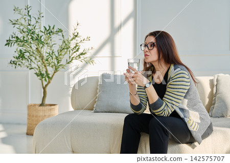 Mature, serious woman with glass of water sitting on sofa, profile view 142575707