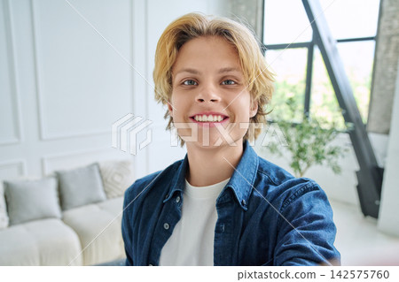 Happy teenage boy smiling at camera while taking selfie in bright modern room Happy teenage boy smiling at camera while taking selfie in bright modern room 142575760