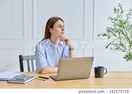 Thoughtful young woman in blue shirt sitting at desk with laptop and looking away Thoughtful young woman in blue shirt sitting at desk with laptop and looking away 142575778
