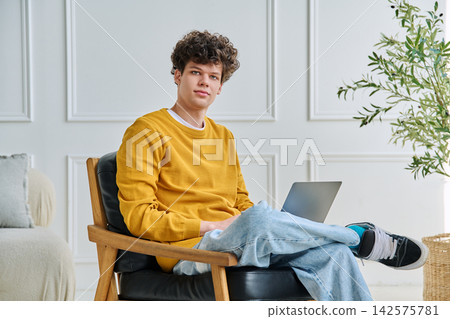 Young man with curly hair sitting in armchair with laptop and looking at camera Young man with curly hair sitting in armchair with laptop and looking at camera 142575781