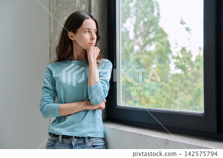 Thoughtful teen girl holding chin and looking out window Thoughtful teen girl holding chin and looking out window 142575794