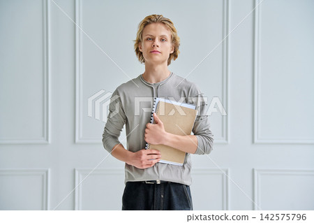 Serious teenage boy holding notebooks and textbooks looking at camera Serious teenage boy holding notebooks and textbooks looking at camera 142575796