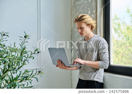 Focused teenage boy working on a laptop near window Focused teenage boy working on a laptop near window 142575806