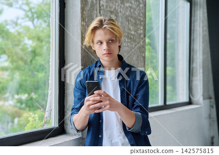 Serious teenage student holding smartphone near window Serious teenage student holding smartphone near window 142575815