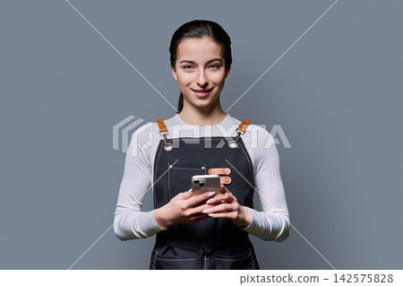 Confident young female in apron using smartphone posing on gray background 142575828