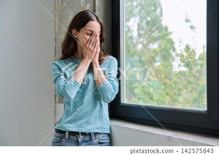 Sad crying teen girl covering her face near window Sad crying teen girl covering her face near window 142575843