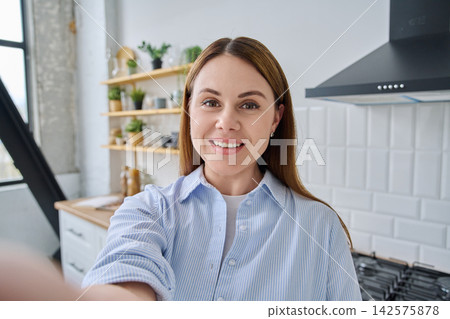 Cheerful woman in blue shirt taking selfie in bright modern kitchen Cheerful woman in blue shirt taking selfie in bright modern kitchen 142575878