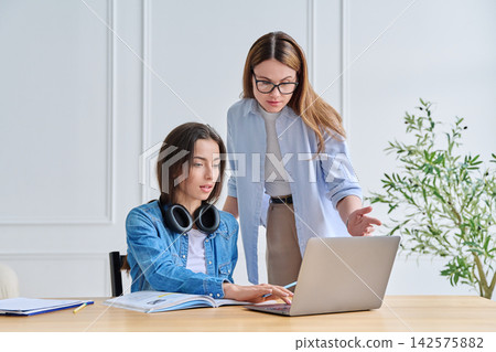 Young female student sitting at desk studying with teacher standing and looking at laptop Young female student sitting at desk studying with teacher standing and looking at laptop 142575882