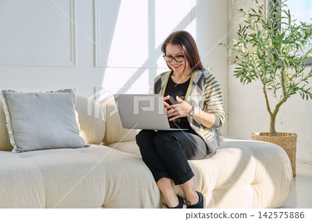 Smiling mature woman having video call on laptop while sitting on sofa in bright living room Smiling mature woman having video call on laptop while sitting on sofa in bright living room 142575886