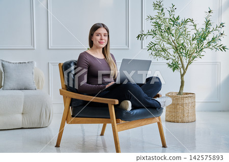 Portrait of young woman sitting on chair with laptop, in home interior Portrait of young woman sitting on chair with laptop, in home interior 142575893