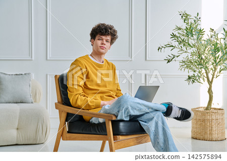 Young man with curly hair sitting in armchair with laptop and looking at camera Young man with curly hair sitting in armchair with laptop and looking at camera 142575894