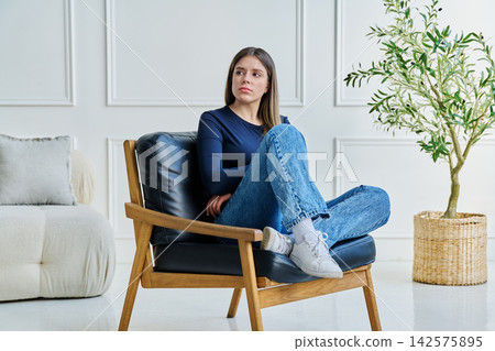 Portrait of thoughtful, pensive young woman sitting in chair at home Portrait of thoughtful, pensive young woman sitting in chair at home 142575895