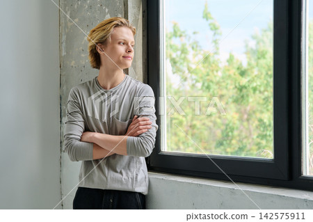 Thoughtful teenage boy with crossed arms by window Thoughtful teenage boy with crossed arms by window 142575911