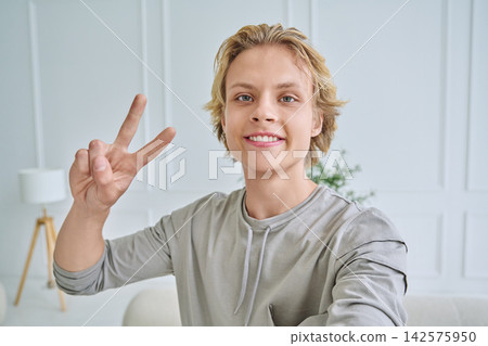 Happy teenage boy smiling at camera and showing peace sign while taking selfie in bright room Happy teenage boy smiling at camera and showing peace sign while taking selfie in bright room 142575950