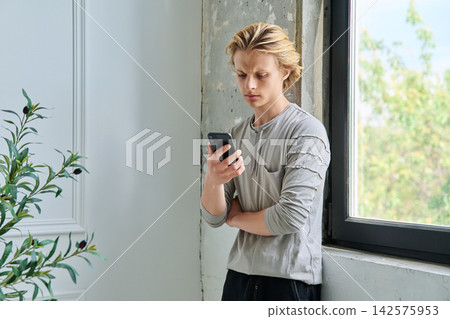 Anxious teenage boy checking smartphone by a window Anxious teenage boy checking smartphone by a window 142575953