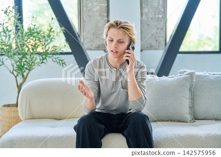 Young blonde man sitting on a comfortable sofa and talking on mobile phone Young blonde man sitting on a comfortable sofa and talking on mobile phone 142575956