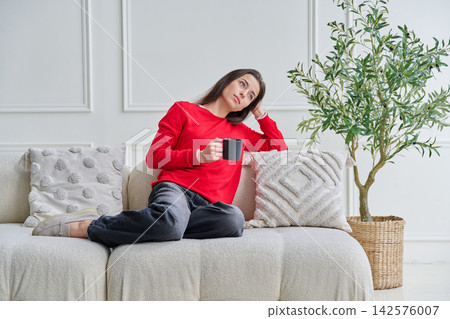 Relaxed, beautiful young woman sitting on couch at home with cup in her hands Relaxed, beautiful young woman sitting on couch at home with cup in her hands 142576007