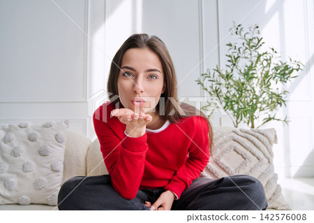 Portrait of beautiful young woman blowing a kiss sitting at home on sofa Portrait of beautiful young woman blowing a kiss sitting at home on sofa 142576008