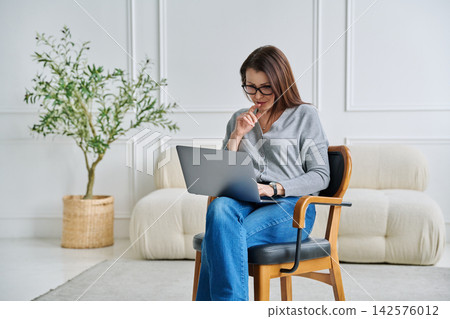 Mature confident female specialist sitting in chair with laptop in home office Mature confident female specialist sitting in chair with laptop in home office 142576012